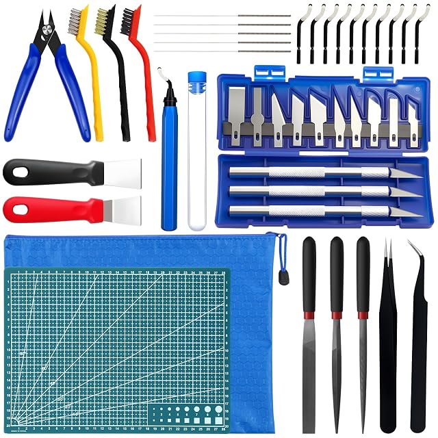 Kit étalé : pince coupante, trois brosses, aiguilles, ébavureur et dix lames, deux grattoirs, un jeu de couteaux dans un étui bleu, un tapis de découpe, une pochette zippée, trois limes et une pince à épiler